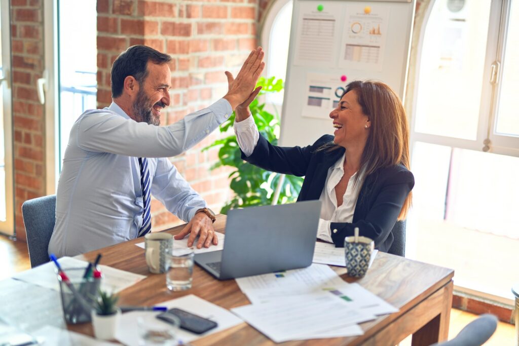 Two people high fiving over an opened laptop. They are sitting at a wooden desk covered in papers