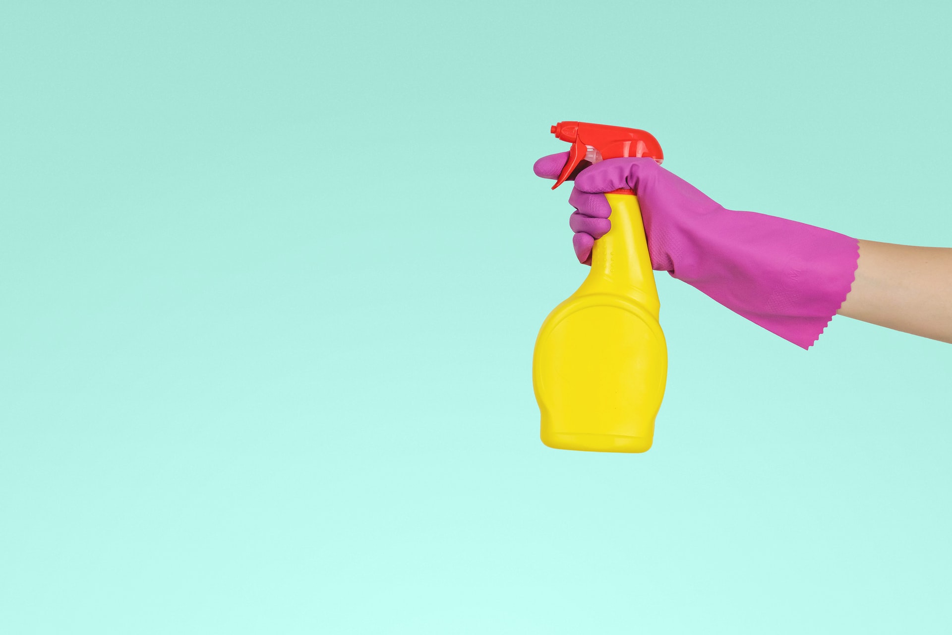 Aqua colored background with a hand wearing a magenta rubber glove, holding a yellow spray bottle