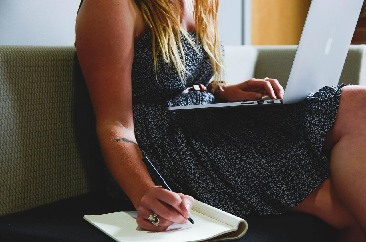 Person sitting on a couch with a laptop on their lap, writing in a notebook with their right hand